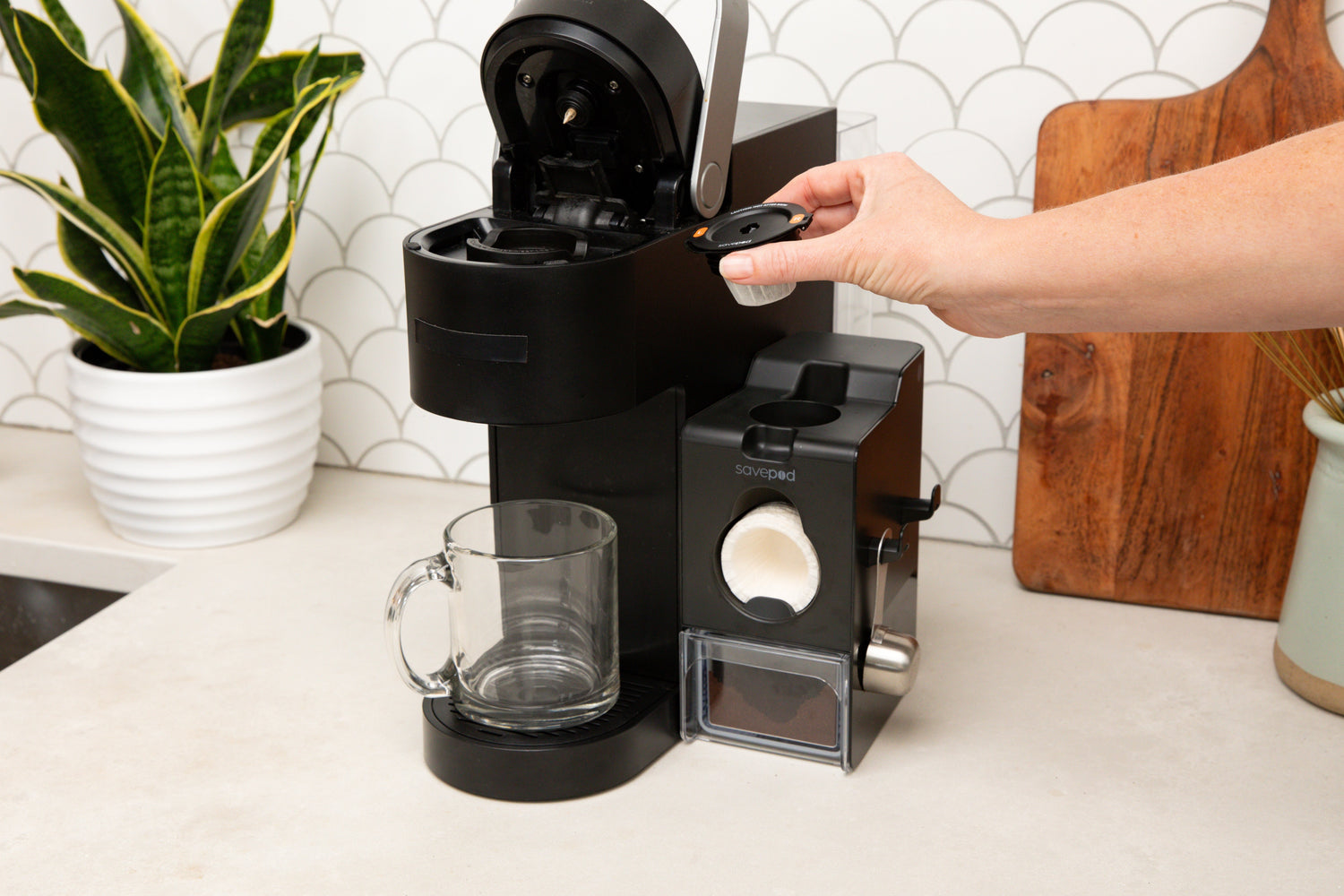 Person using a coffee machine on a kitchen counter with a plant and cutting board in the background.